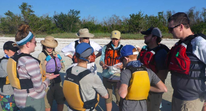 A group of young people and their parents wear life jackets and stand in a circle on a beach.